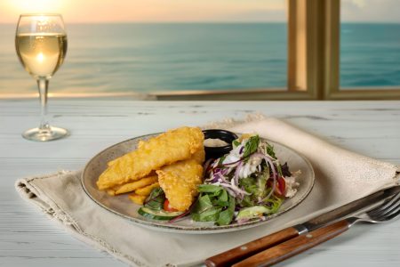 Plate of fish and chips with salad and white wine on a table overlooking the ocean at sunset, photographed in Newcastle.