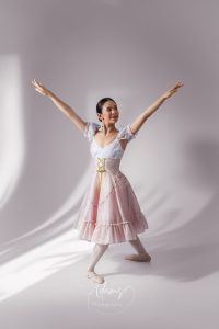 Ballet dancer in a pink and white classical costume posing with arms raised in a softly lit Newcastle studio.