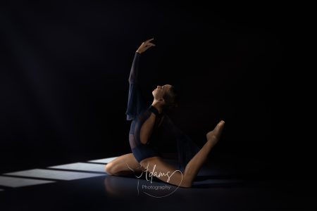 Contemporary dancer in a floor pose with one leg extended and arm raised, lit by soft window‑style light in a Newcastle studio.